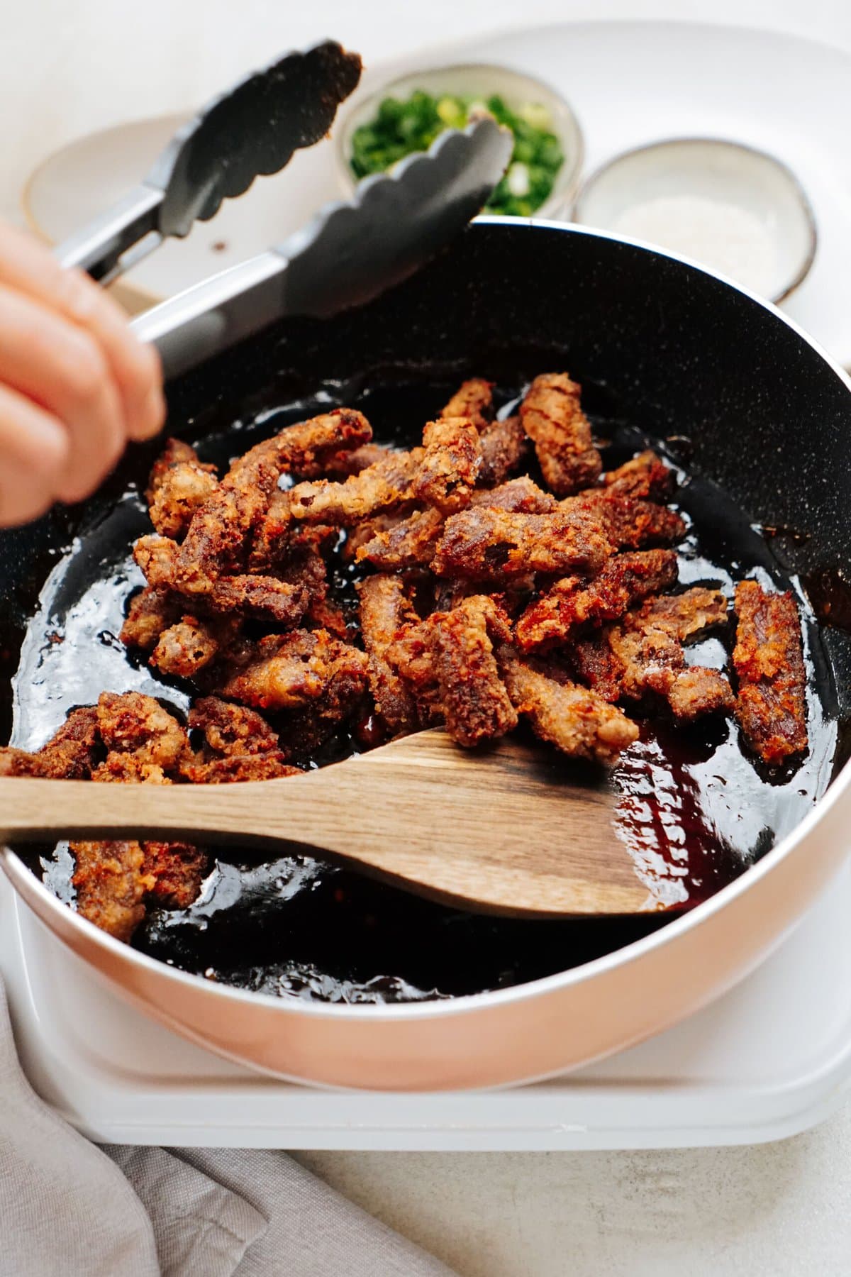 A person uses a wooden spoon to move crispy beef pieces in a black skillet. A wooden spoon rests in the skillet. Bowls of chopped green onions and a white sauce are in the background.