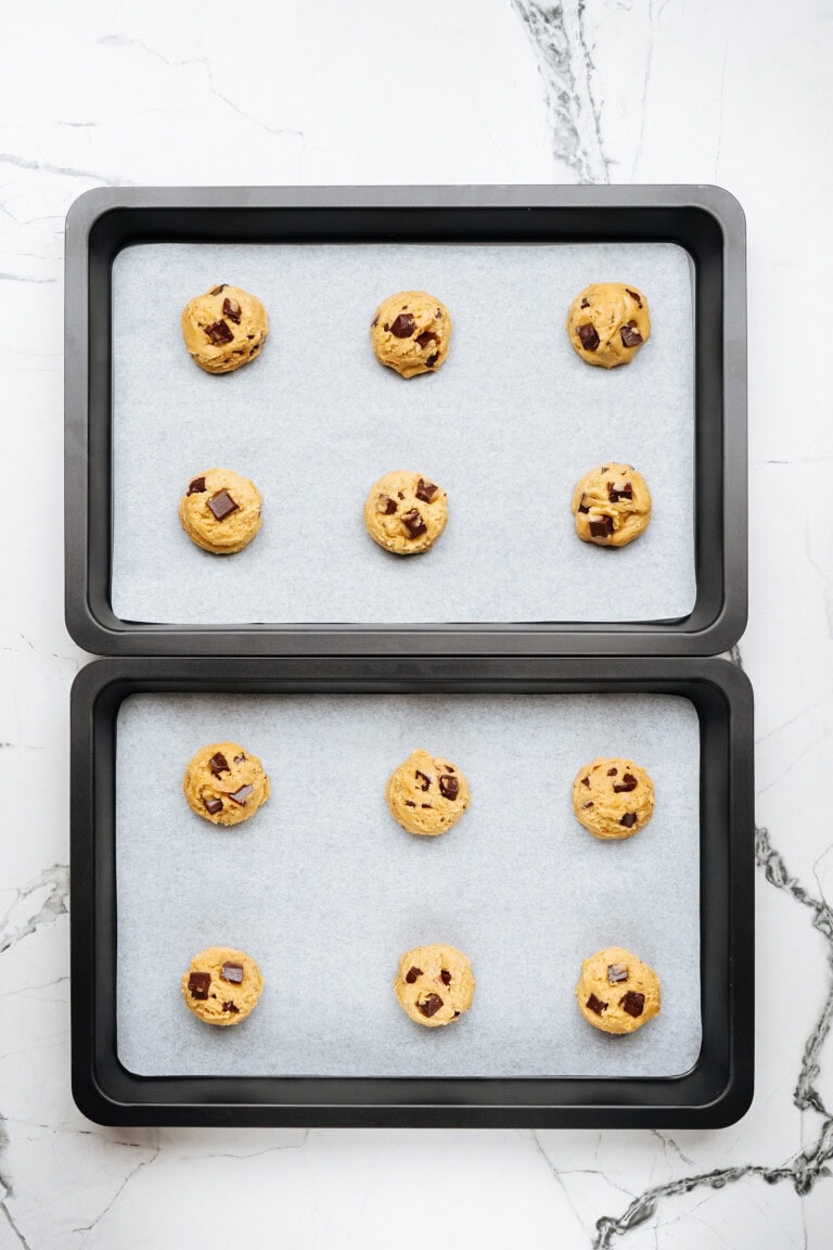 Two baking trays lined with parchment paper hold raw chocolate chip cookie dough balls, ready for baking into delicious chocolate chip cookies.