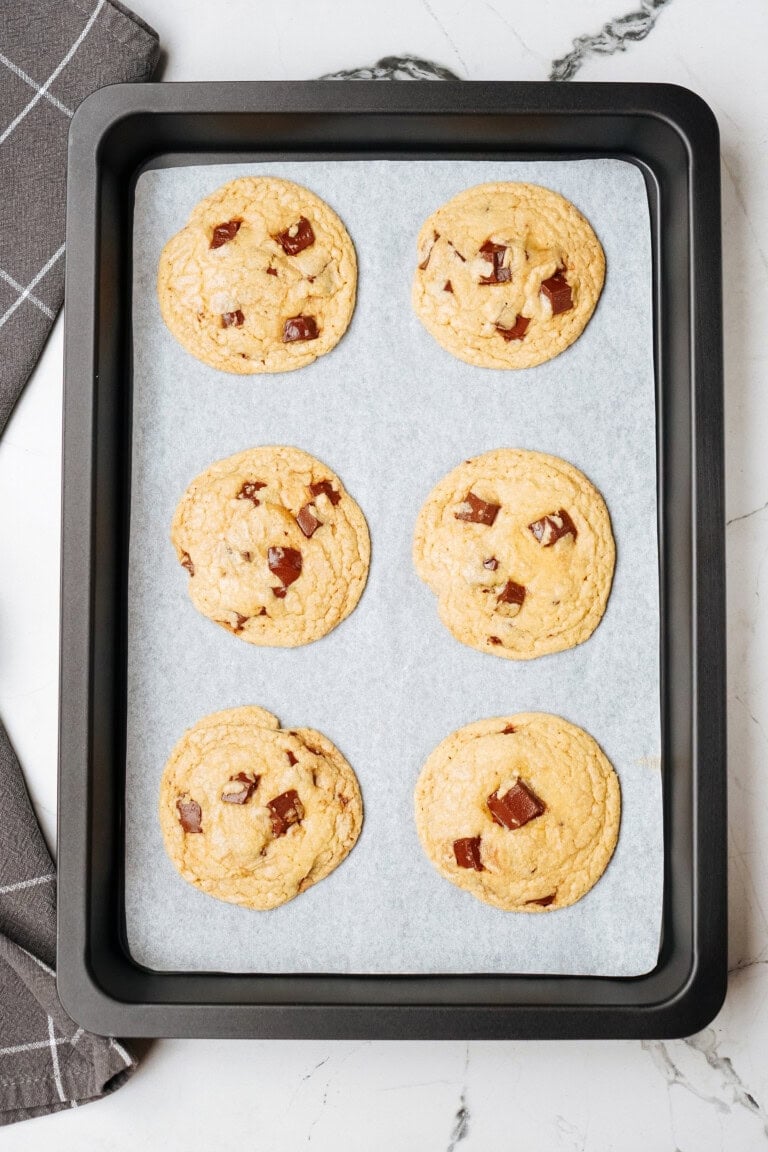 A baking tray with six freshly baked chocolate chip cookies rests on parchment paper, placed on a marble countertop next to a folded gray kitchen towel.