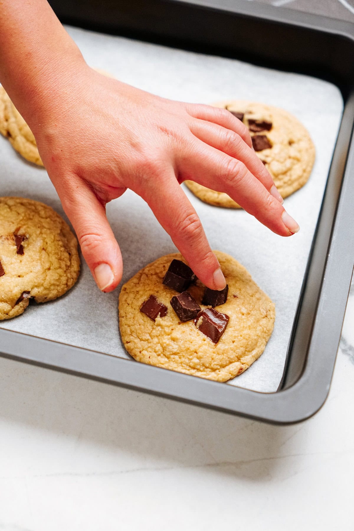 A hand placing chocolate chip cookies on a baking sheet.