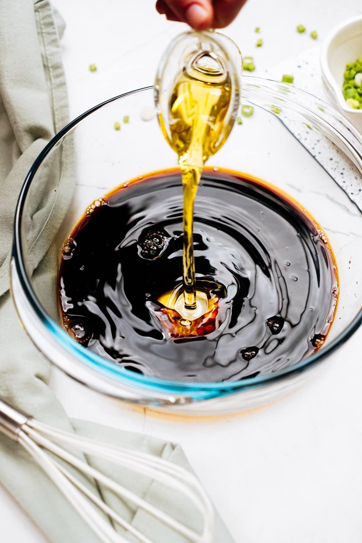 A hand pouring oil from a small glass pitcher into a bowl containing a dark liquid, with a whisk, green cloth, and chopped green onions nearby—perfect ingredients for marinating Korean chicken.