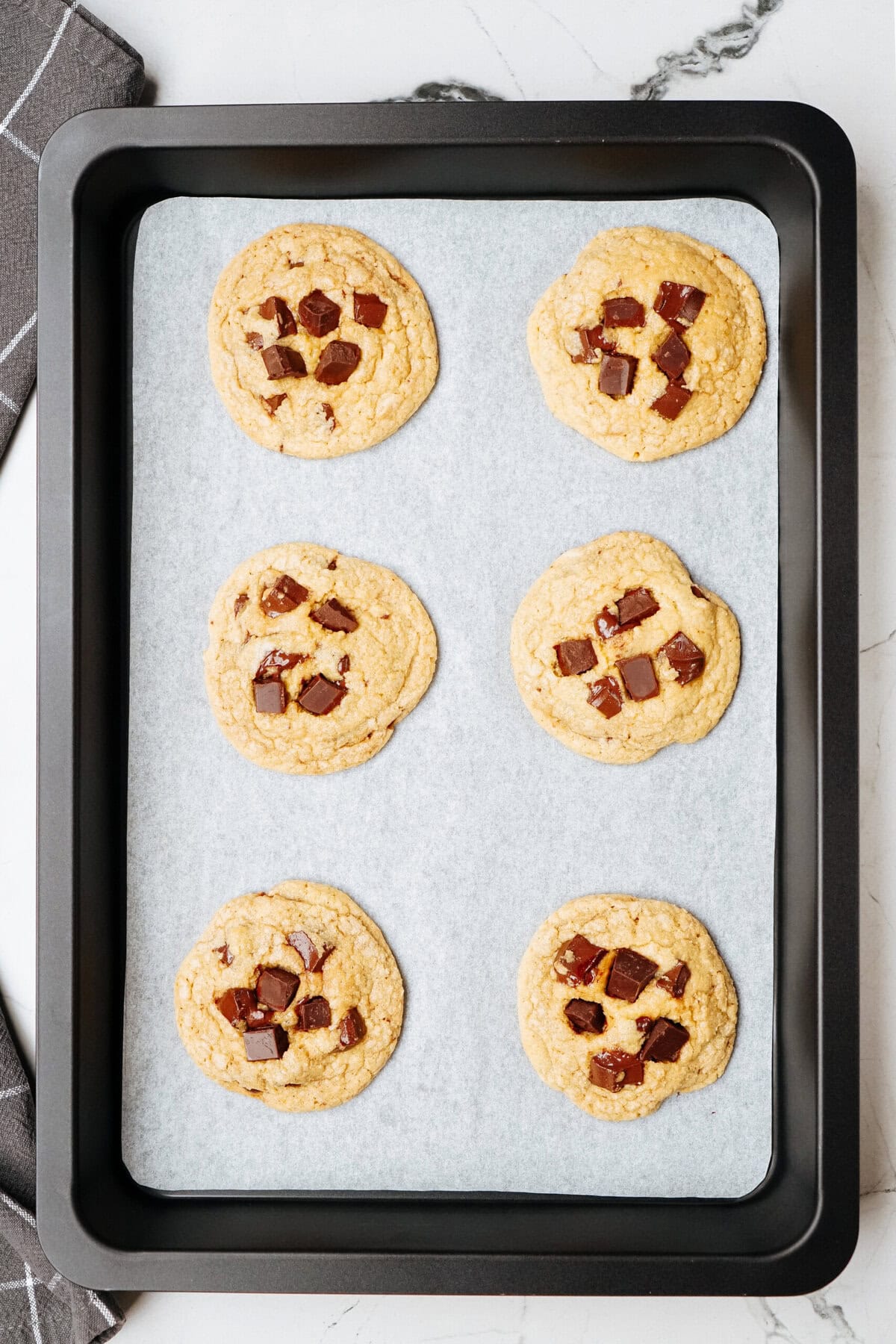 A black baking tray holds six delicious chocolate chip cookies on a sheet of parchment paper.