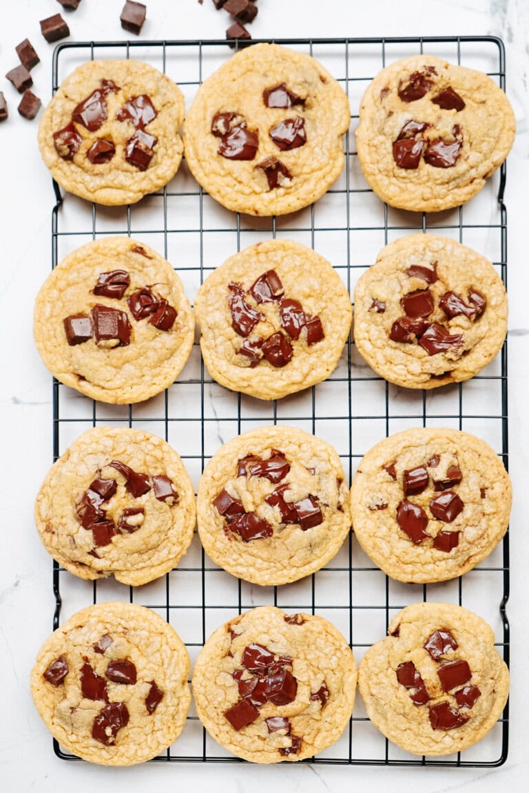 Twelve delicious chocolate chip cookies cooling on a black wire rack with a few extra chocolate chunks scattered on the white surface.