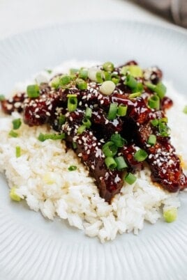 A dish of white rice topped with glazed meat strips, sprinkled with sesame seeds and chopped green onions.