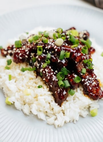 A dish of white rice topped with glazed meat strips, sprinkled with sesame seeds and chopped green onions.