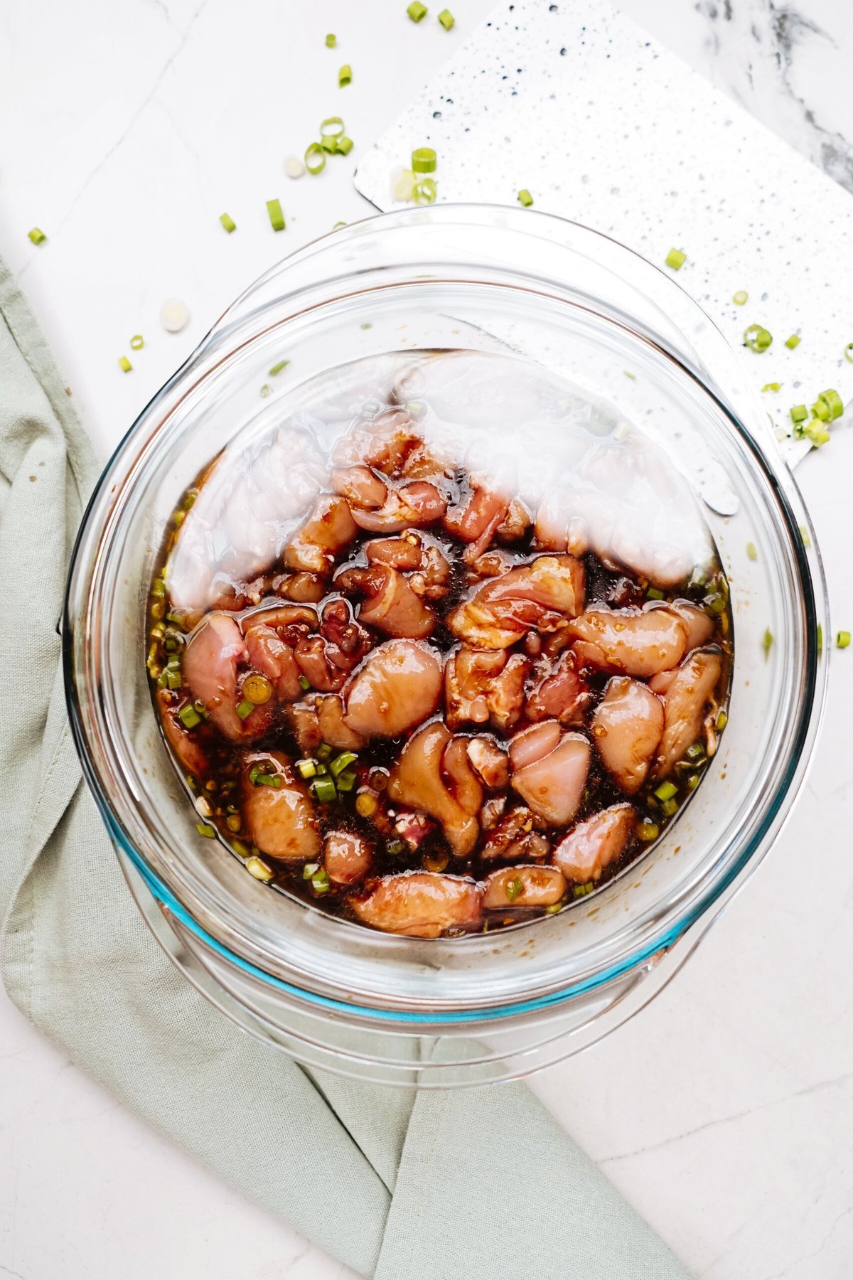 A glass bowl filled with marinated Korean chicken pieces, topped with chopped green onions, sits on a light-colored surface next to a green cloth napkin.