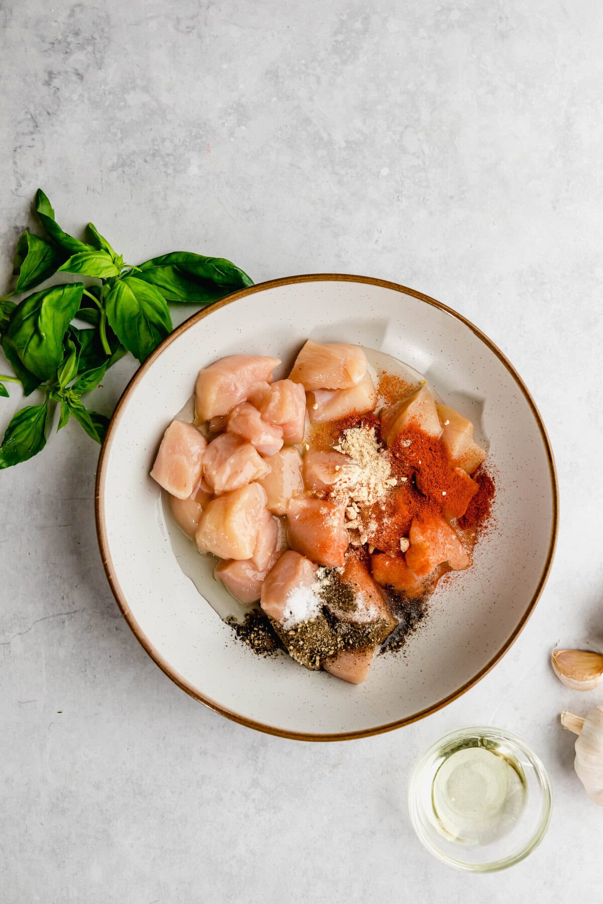 A bowl of raw chopped chicken seasoned with various spices next to basil leaves, garlic cloves, and a small container of oil.