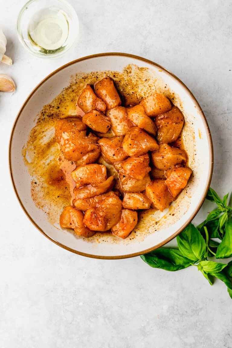 A white bowl containing seasoned, raw chicken chunks is placed on a light surface. Fresh basil leaves and a small glass bowl with oil are nearby.