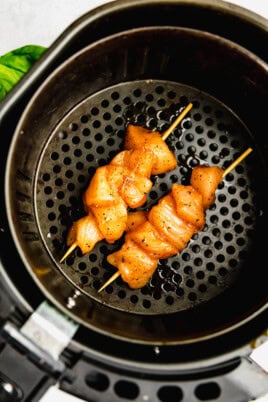 Two raw, seasoned chicken skewers placed in an air fryer basket ready for cooking.