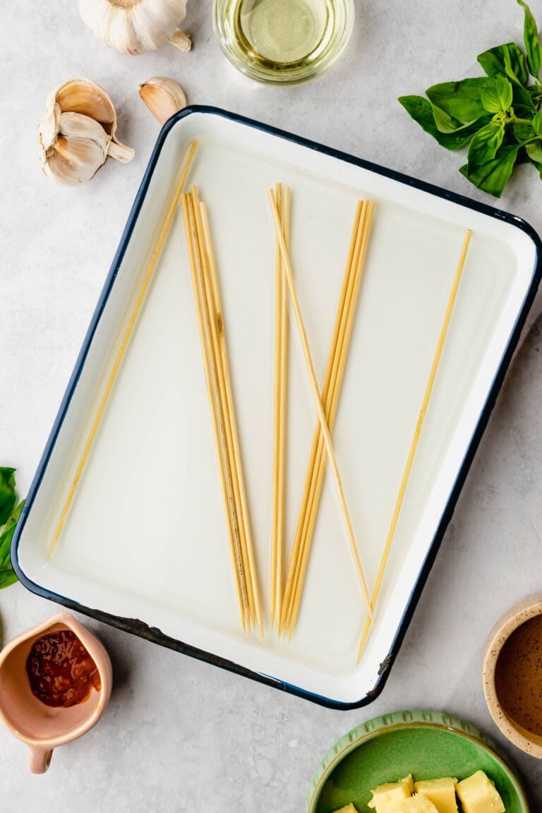 A white enamel tray contains several wooden skewers soaking in water. Surrounding the tray are various ingredients, including garlic, basil leaves, oil, and sauces.