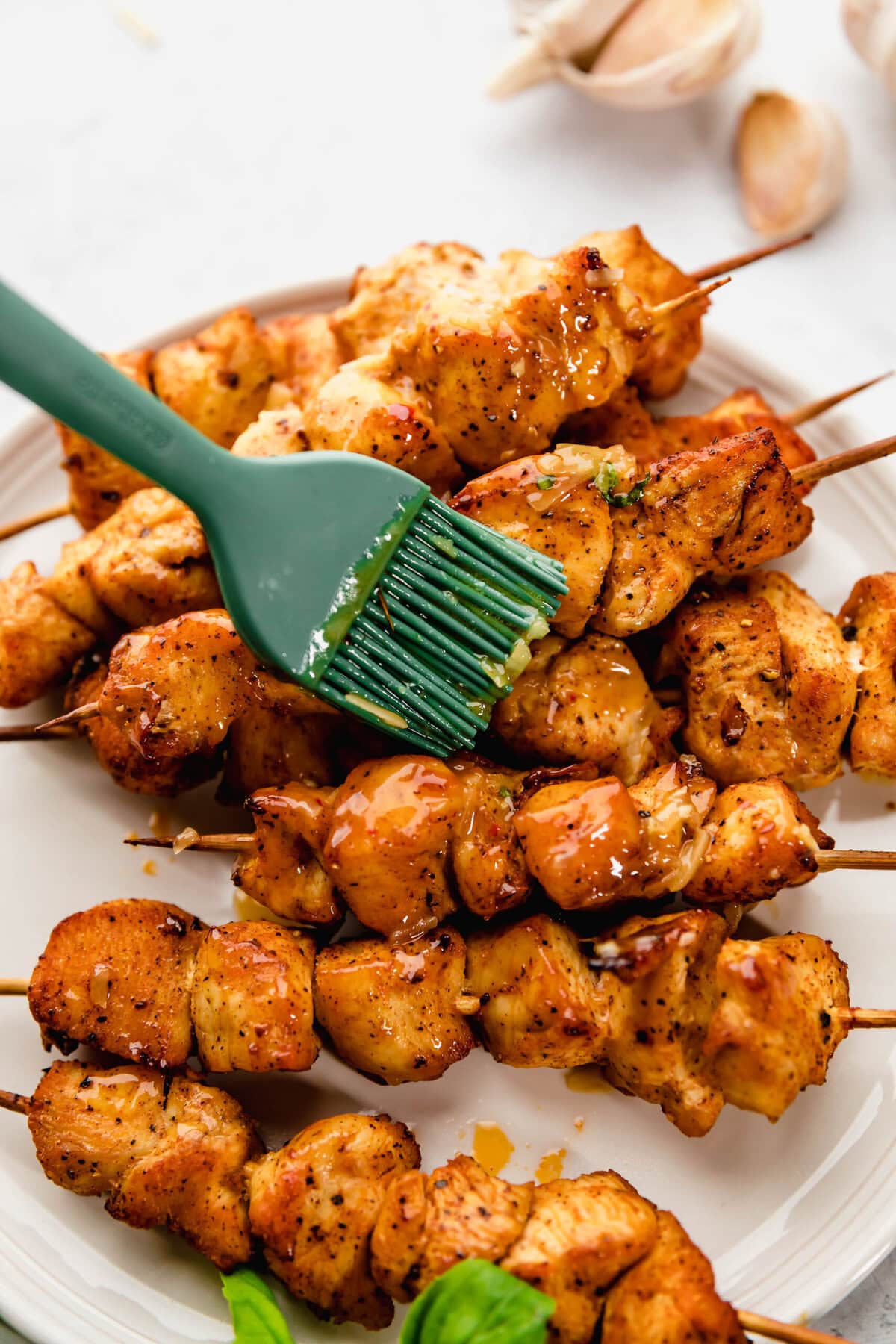 A green brush applies glaze to cooked chicken skewers on a white plate, with garlic cloves and basil leaves in the background.