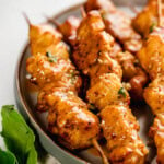 Grilled chicken skewers coated in a glaze are arranged on a round plate. A sprig of basil is placed beside the plate.