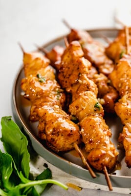 Grilled chicken skewers coated in a glaze are arranged on a round plate. A sprig of basil is placed beside the plate.