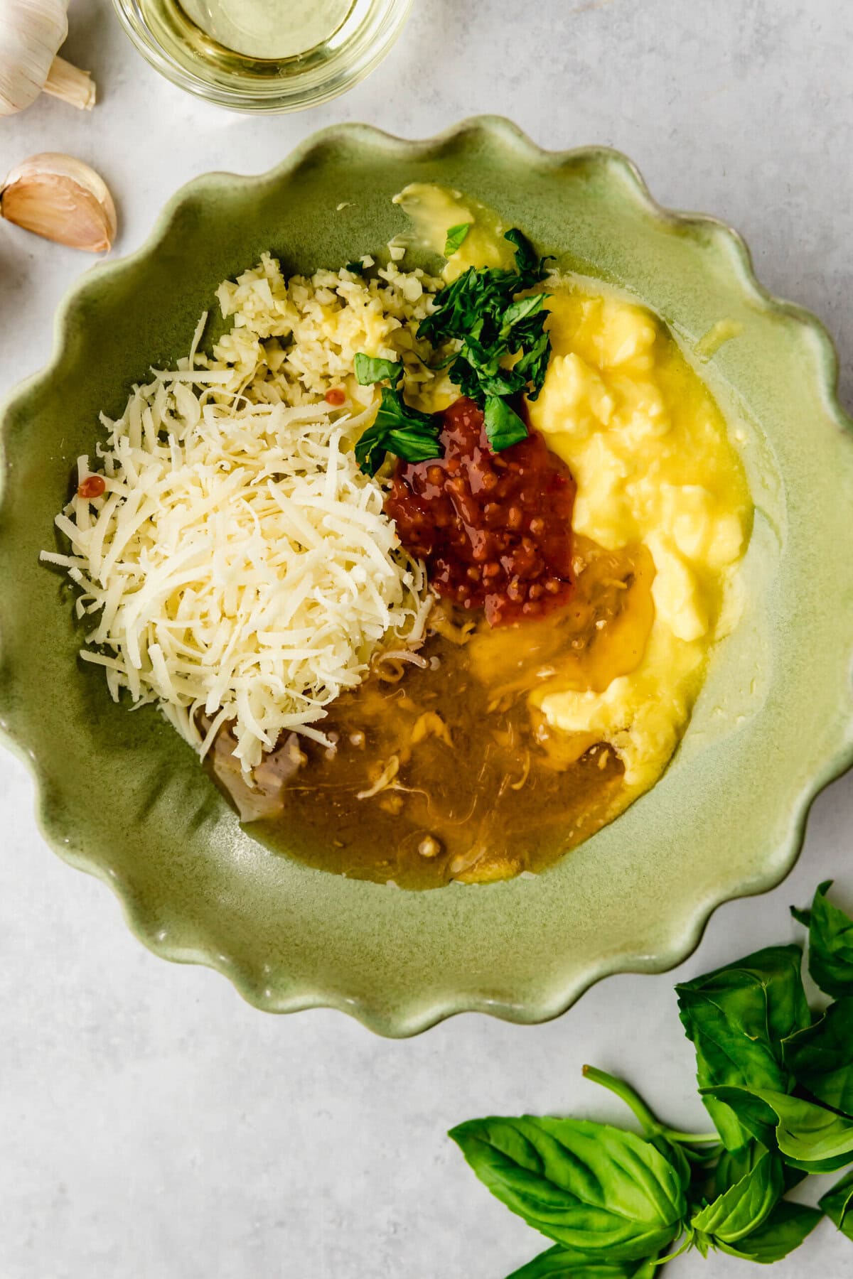 A green bowl contains portions of scrambled eggs, shredded cheese, tomato salsa, sautéed spinach, and crumbled feta cheese. Fresh basil leaves and garlic cloves are nearby.