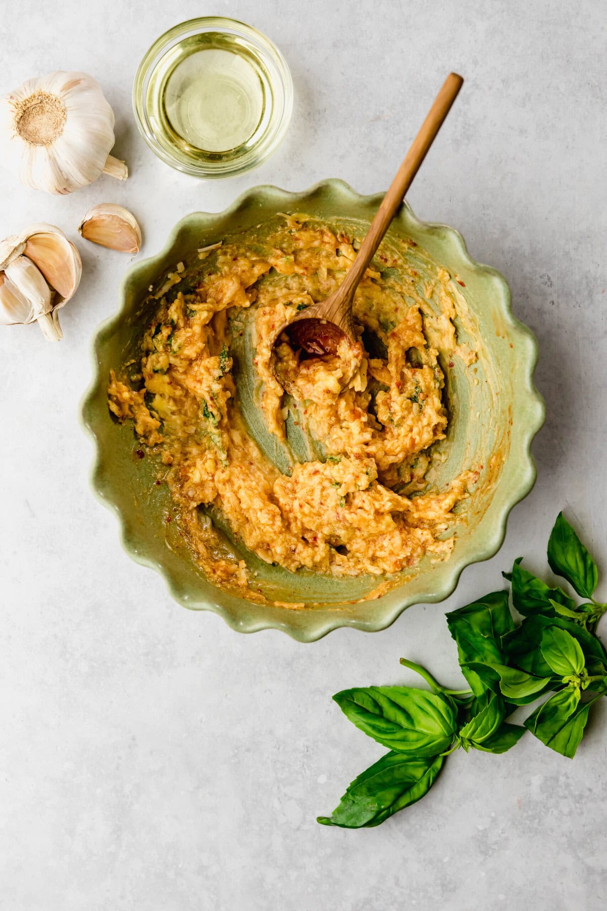 A green bowl filled with a thick, yellowish mixture, a wooden spoon, garlic cloves, a small dish of oil, and fresh basil leaves are arranged on a light gray surface.