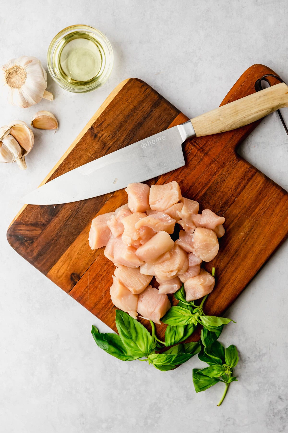 Diced raw chicken on a wooden cutting board with a knife, garlic cloves, a small dish of oil, and fresh basil leaves on a light gray surface.