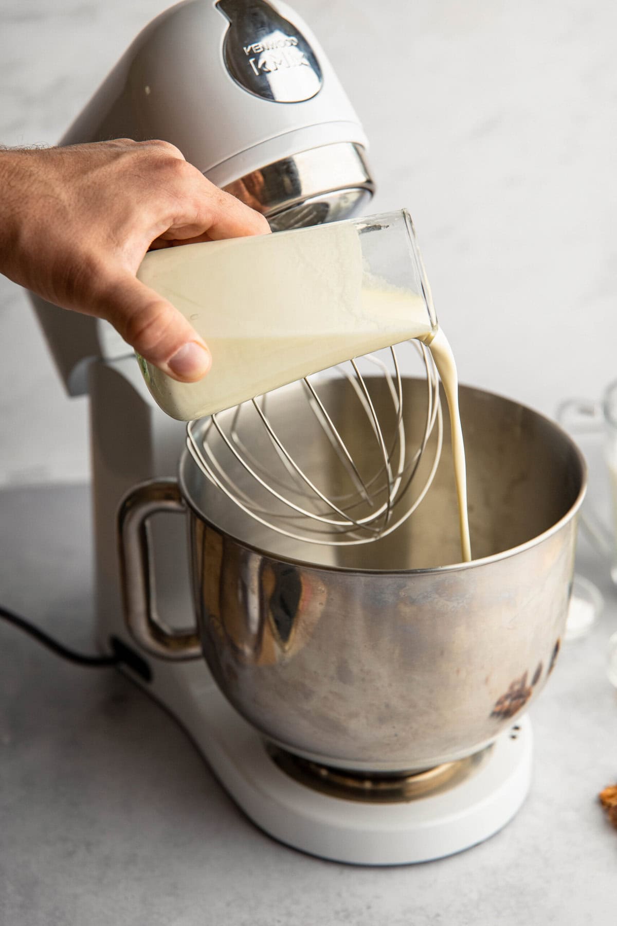 A person pours a creamy liquid from a glass bottle into the bowl of a stand mixer with a whisk attachment.