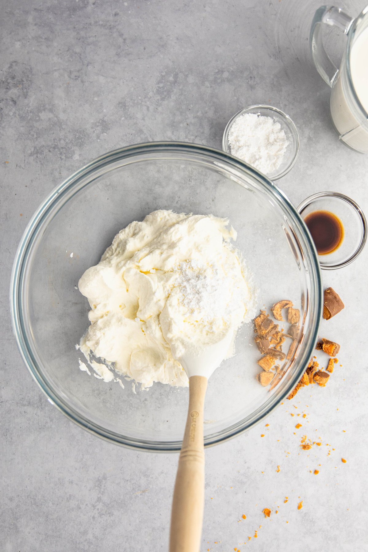 A glass bowl containing whipped cream, powdered sugar, vanilla extract, and crushed toffee bits on a gray surface. A wooden spoon rests inside the bowl.