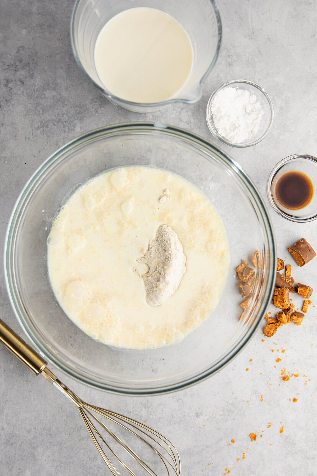 A glass bowl with milk and dry ingredients, surrounded by a whisk, broken cookie pieces, a measuring cup of milk, a small bowl of flour, and a small bowl of vanilla extract on a gray surface.