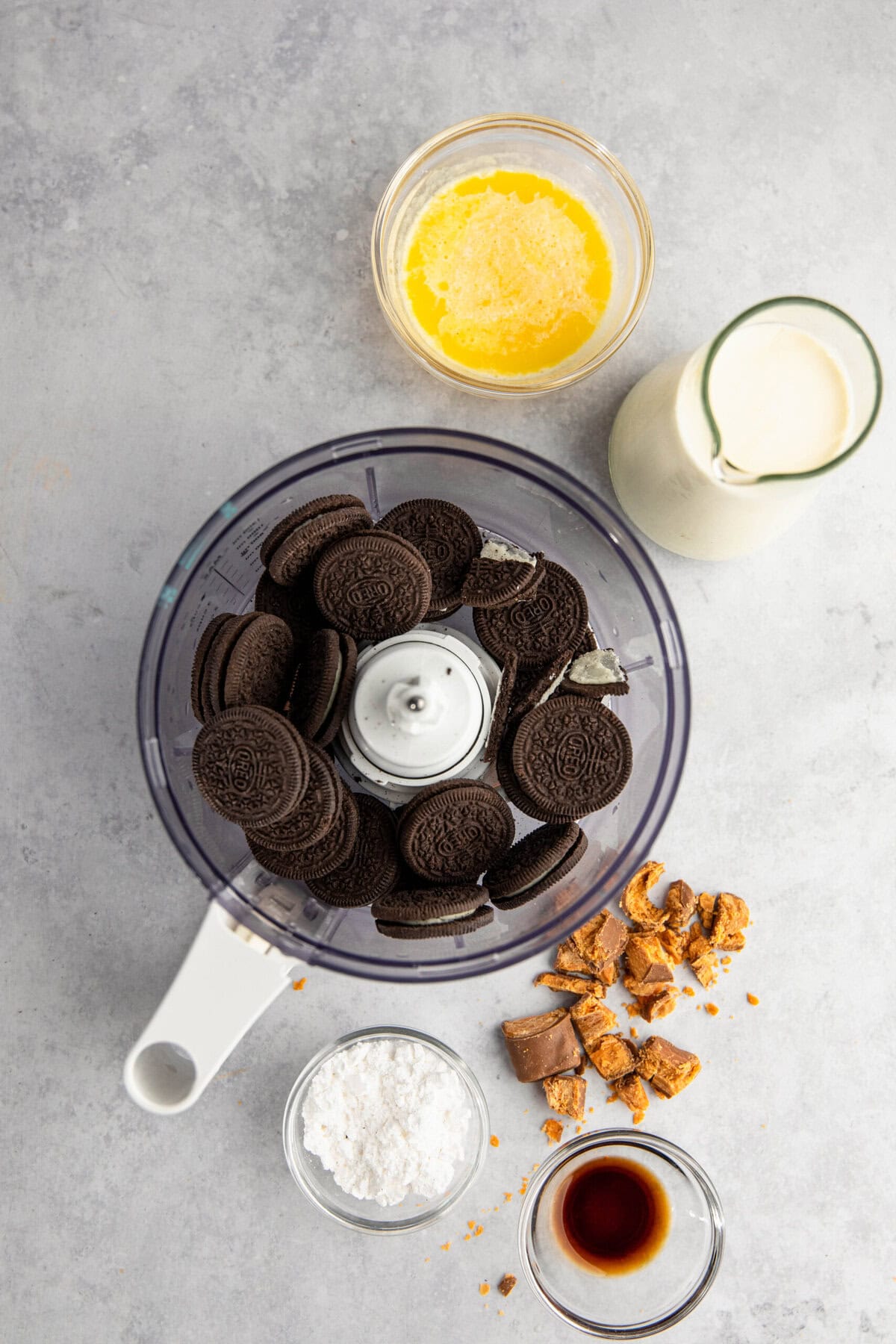 A food processor bowl filled with chocolate sandwich cookies, surrounded by bowls containing melted butter, cream, powdered sugar, broken cookies, and vanilla extract on a light gray surface.