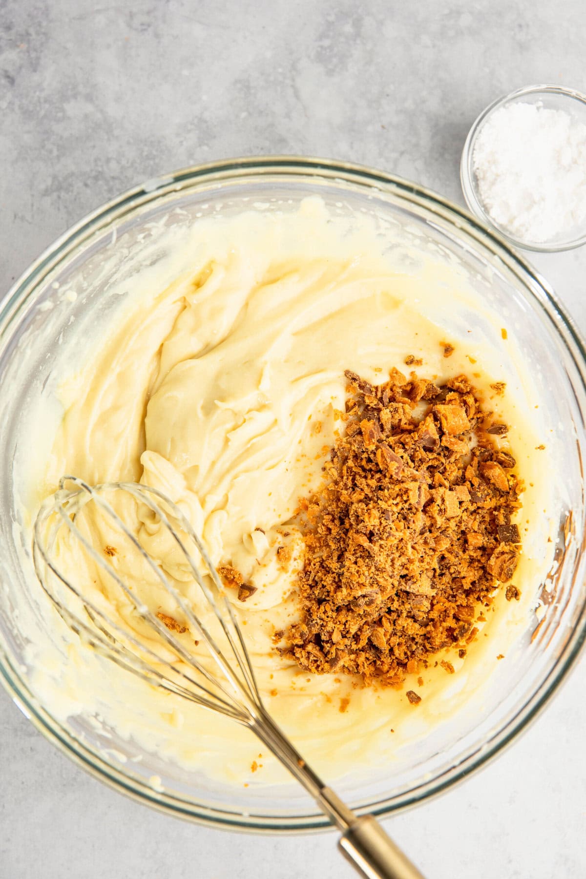 A mixing bowl with creamy yellow batter and a topping of crumbled brown pieces, beside a whisk and a small bowl of white powder on a gray surface.