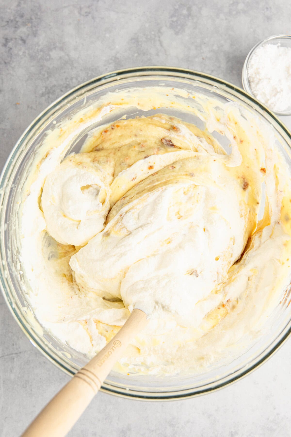 A glass bowl with whipped cream and banana slices, being mixed with a wooden spoon. A small bowl of flour is visible in the background.