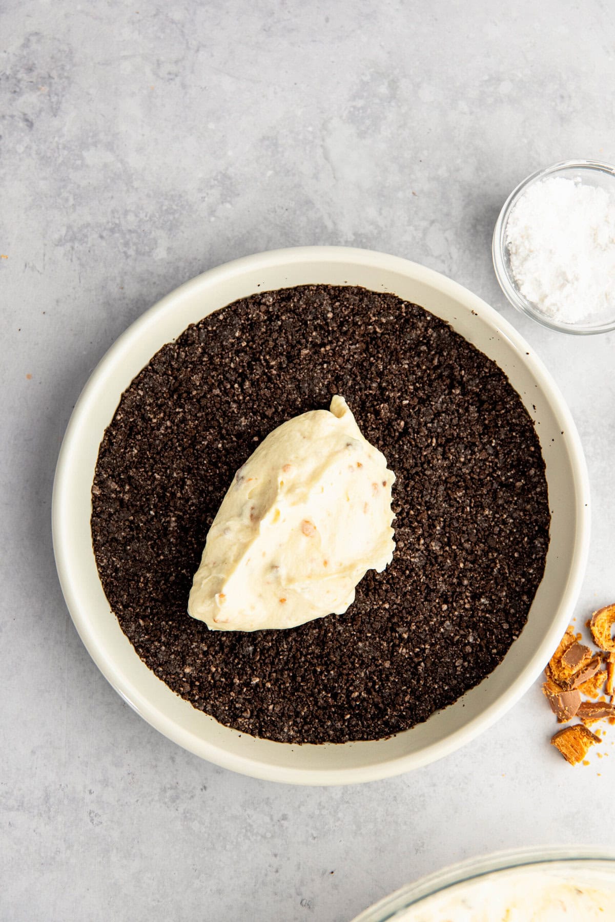 A pie dish filled with crushed chocolate cookie base and topped with a dollop of creamy filling. To the side, there is a small bowl of salt and some crushed candy pieces on the gray surface.