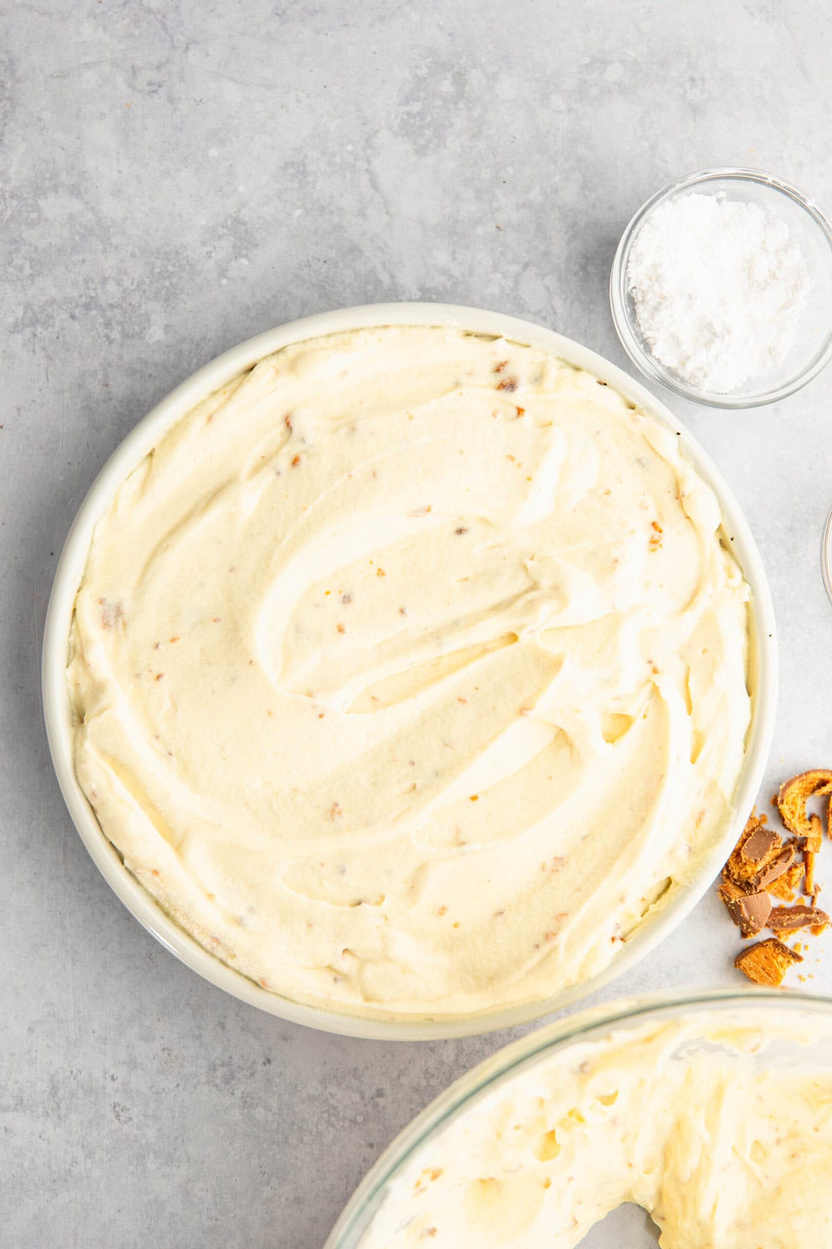 A bowl filled with creamy, light-colored dessert mixture on a gray surface, with a small glass bowl of white powder and a few crumbled brown pieces nearby.