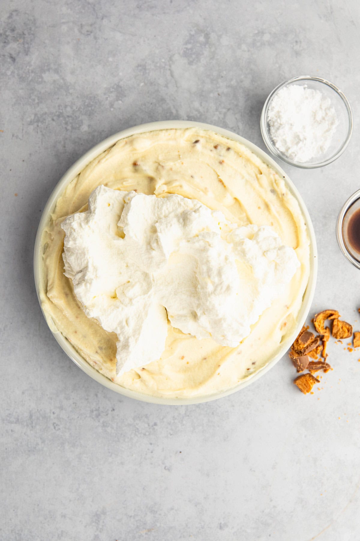 A bowl of creamy dessert with a dollop of whipped cream on top, accompanied by small bowls of powdered sugar and syrup, and some scattered caramel pieces on a light gray surface.