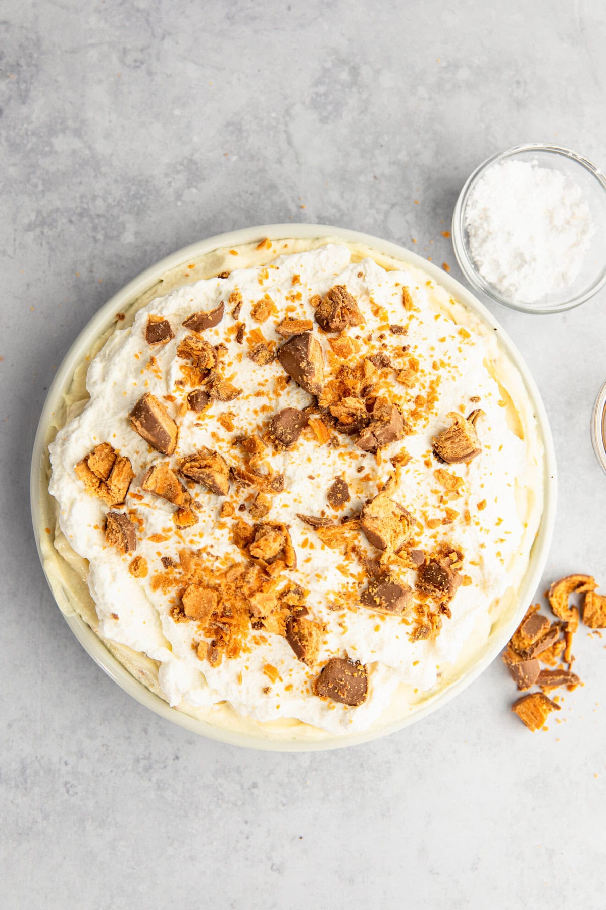 A pie topped with whipped cream and crumbled pieces of a caramel and chocolate candy bar, placed on a grey surface with a small bowl of powdered sugar nearby.