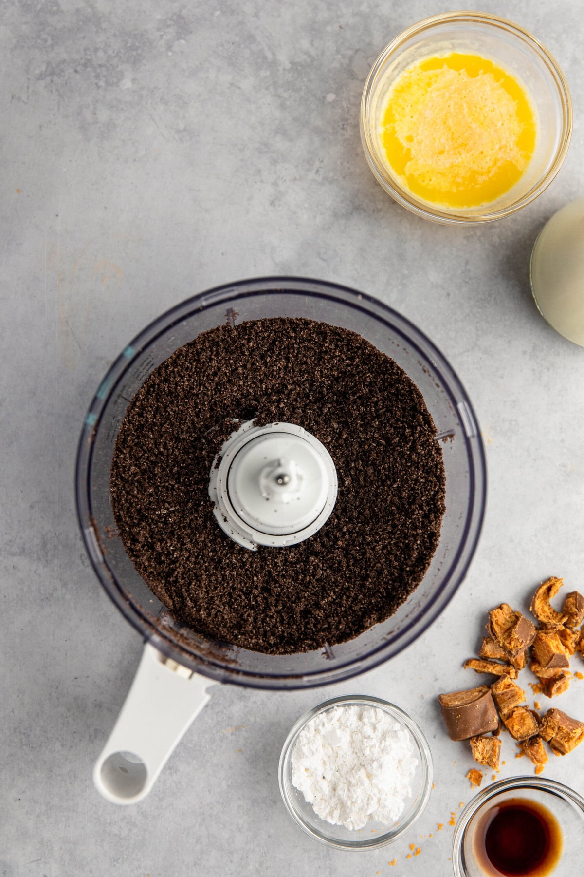 Ingredients for a recipe are laid out on a counter: a food processor with ground chocolate cookies, a bowl of melted butter, two small bowls of powdered sugar and vanilla extract, and chopped nuts.