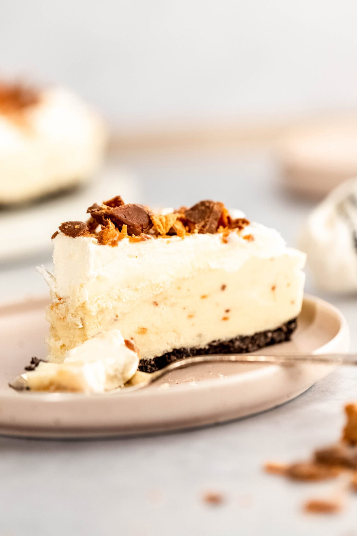 A slice of creamy pie with a chocolate crust, topped with whipped cream and crumbled pieces, is on a beige plate with a forkful of pie next to it.