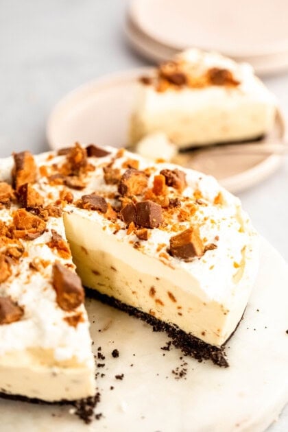 A creamy pie with a black crust, topped with whipped cream and crumbled pieces of candy, with a slice missing. Two plates of pie are in the background.