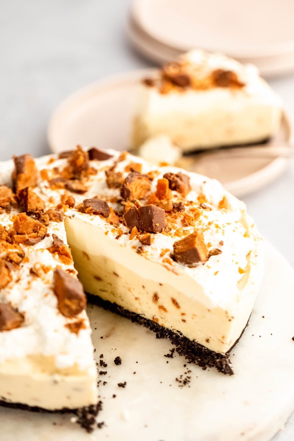 A creamy pie with a black crust, topped with whipped cream and crumbled pieces of candy, with a slice missing. Two plates of pie are in the background.