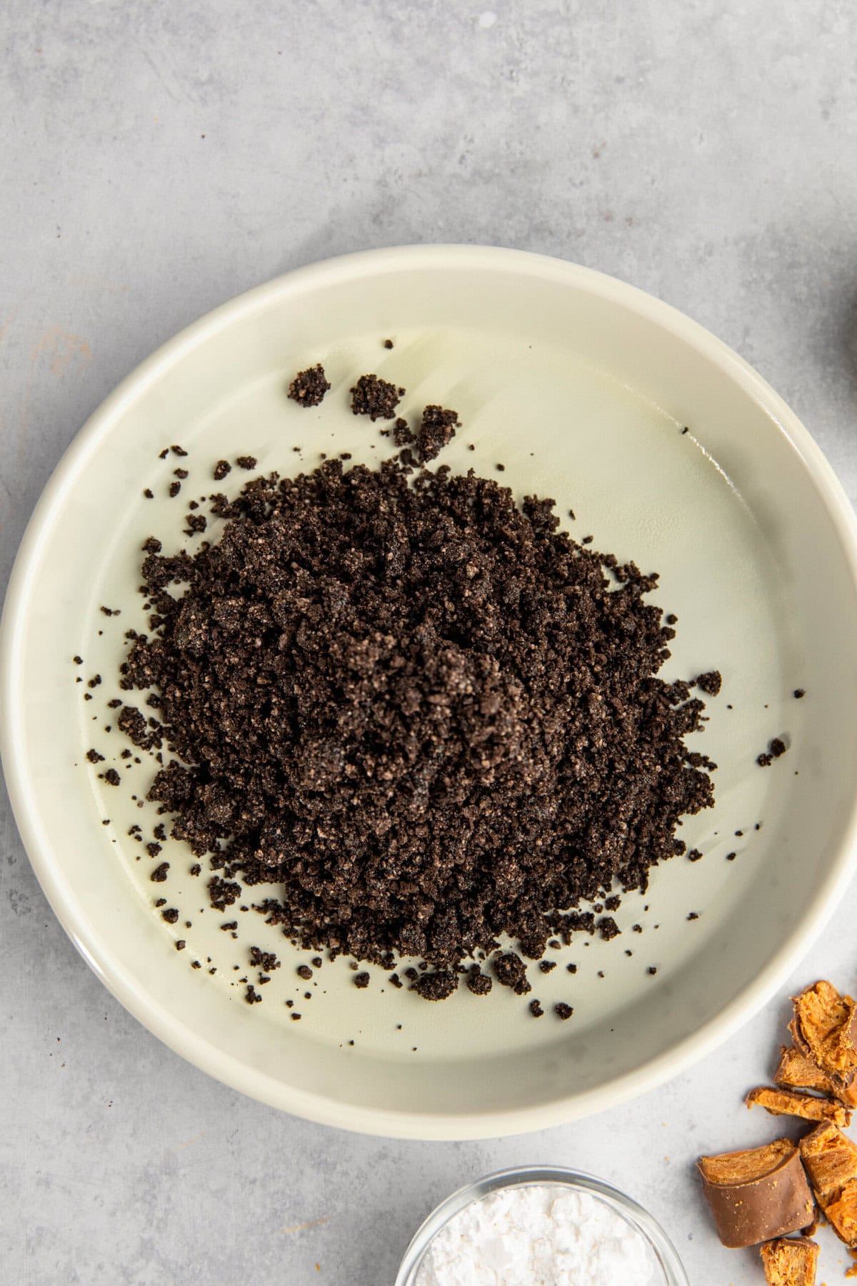 A bowl contains a pile of crumbly, dark brown substance on a light-colored surface. Small portions of other ingredients are partially visible around the bowl.