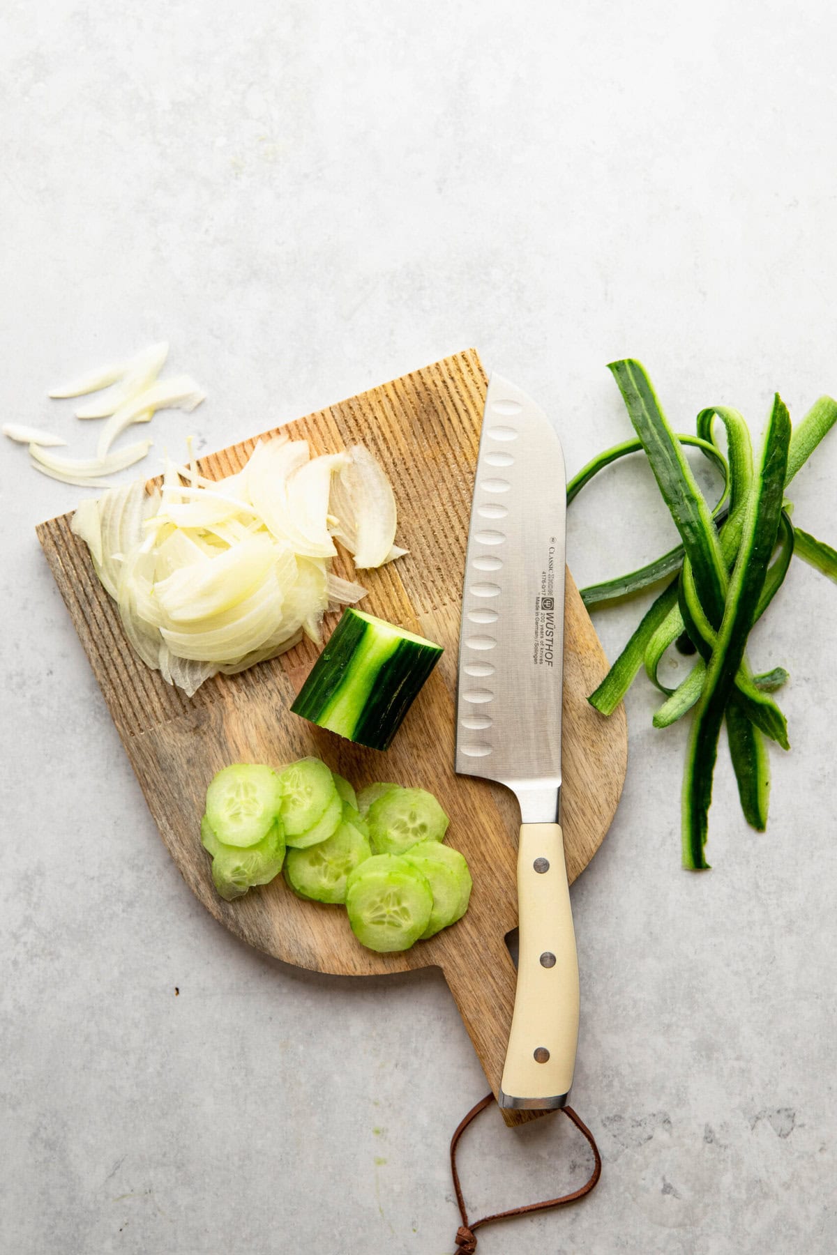 A cutting board with sliced cucumber, shredded onion, and green vegetable slices, accompanied by a knife.