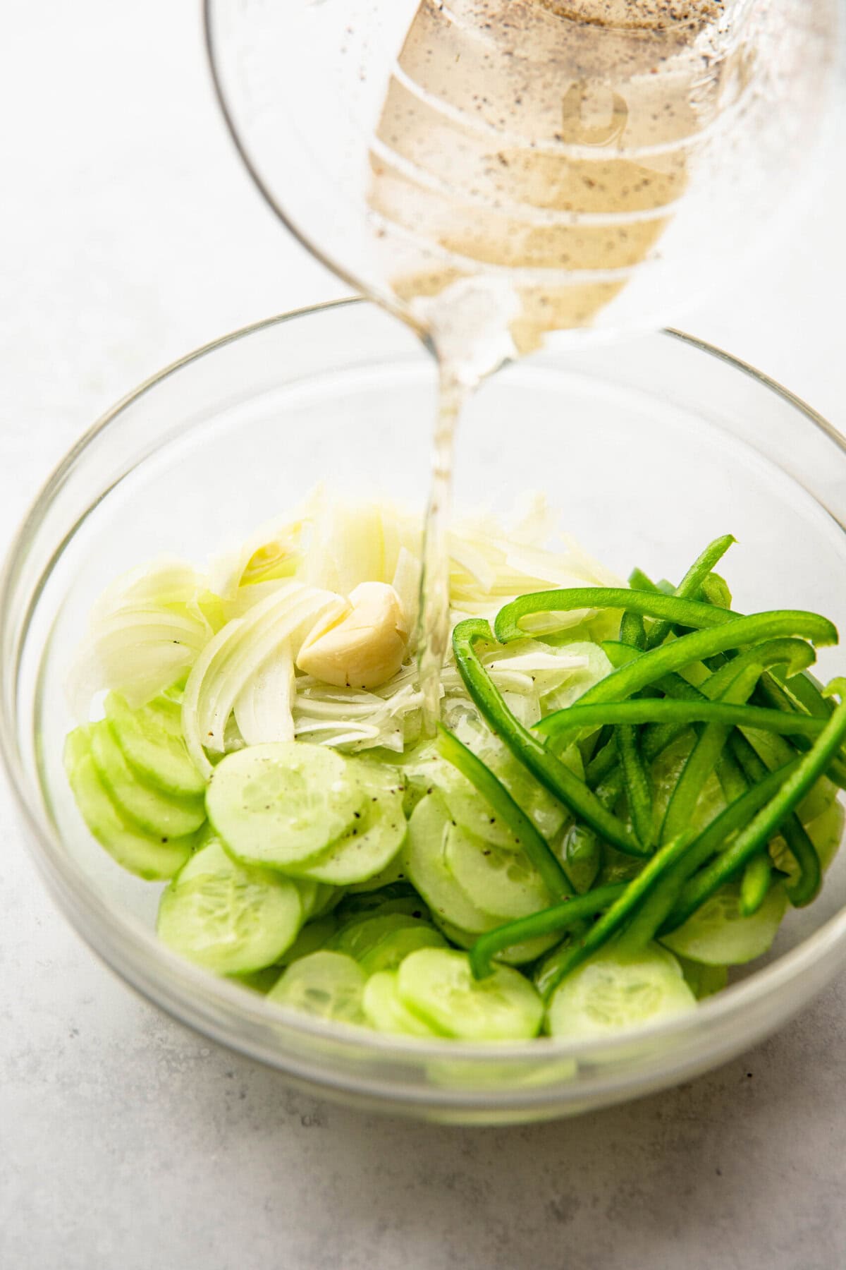 A clear glass bowl contains sliced cucumbers, green bell peppers, and onions, with dressing being poured over the vegetables from a measuring cup.