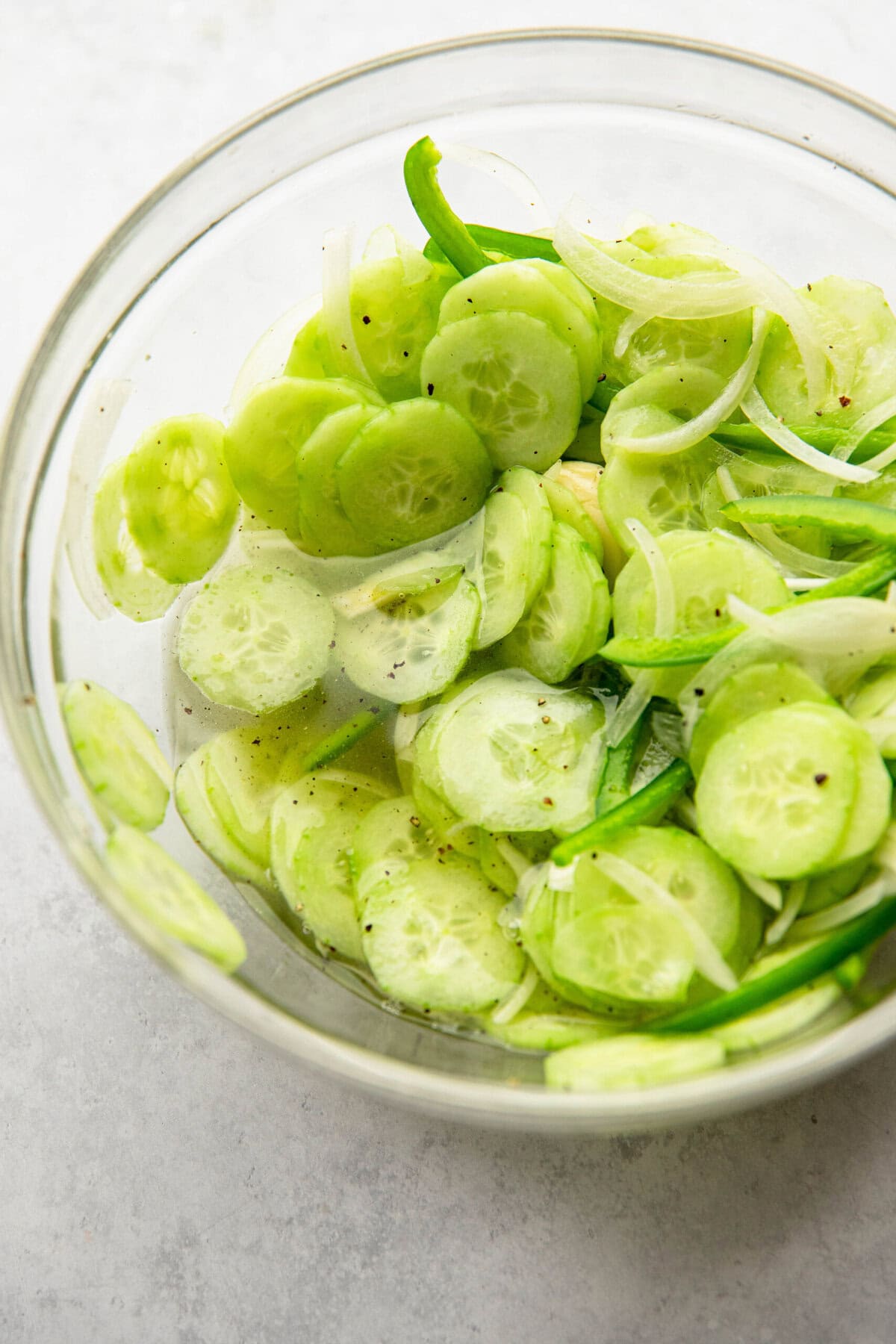 A clear glass bowl containing sliced cucumbers, onions, and green peppers, seasoned with black pepper and mixed in a light liquid.