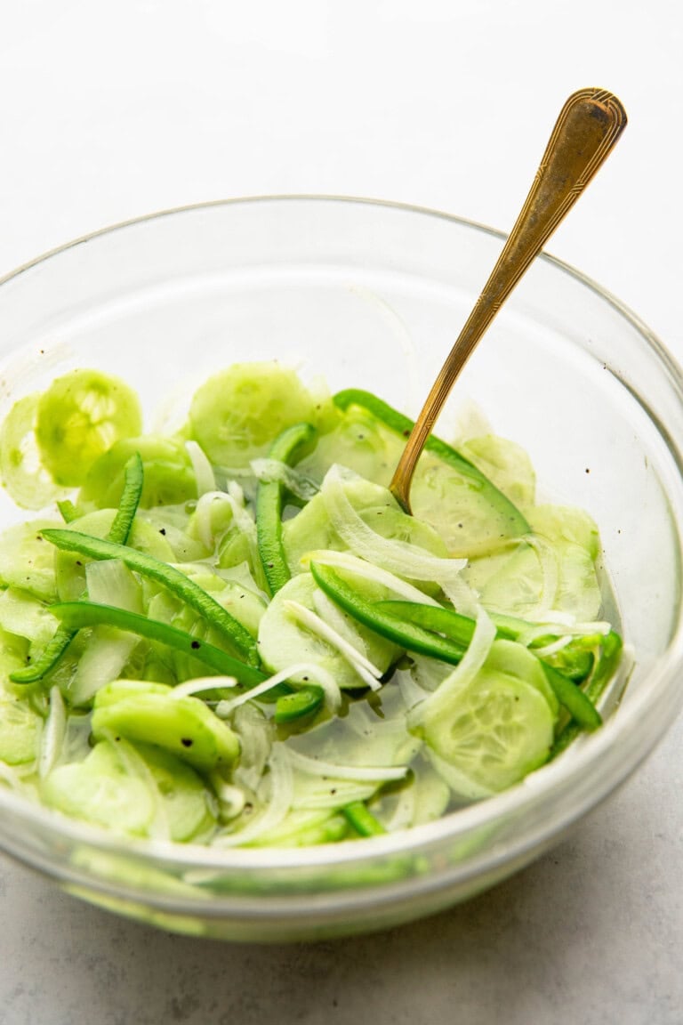 A glass bowl containing sliced cucumbers, green bell peppers, and onions, with a fork resting inside.