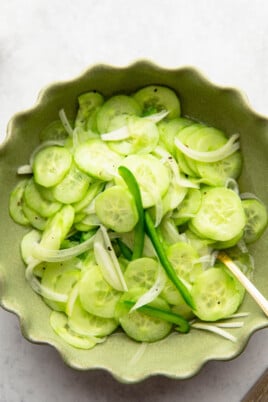 A green bowl filled with sliced cucumbers, thinly sliced onions, and green chili pepper pieces. A silver spoon is resting inside the bowl.