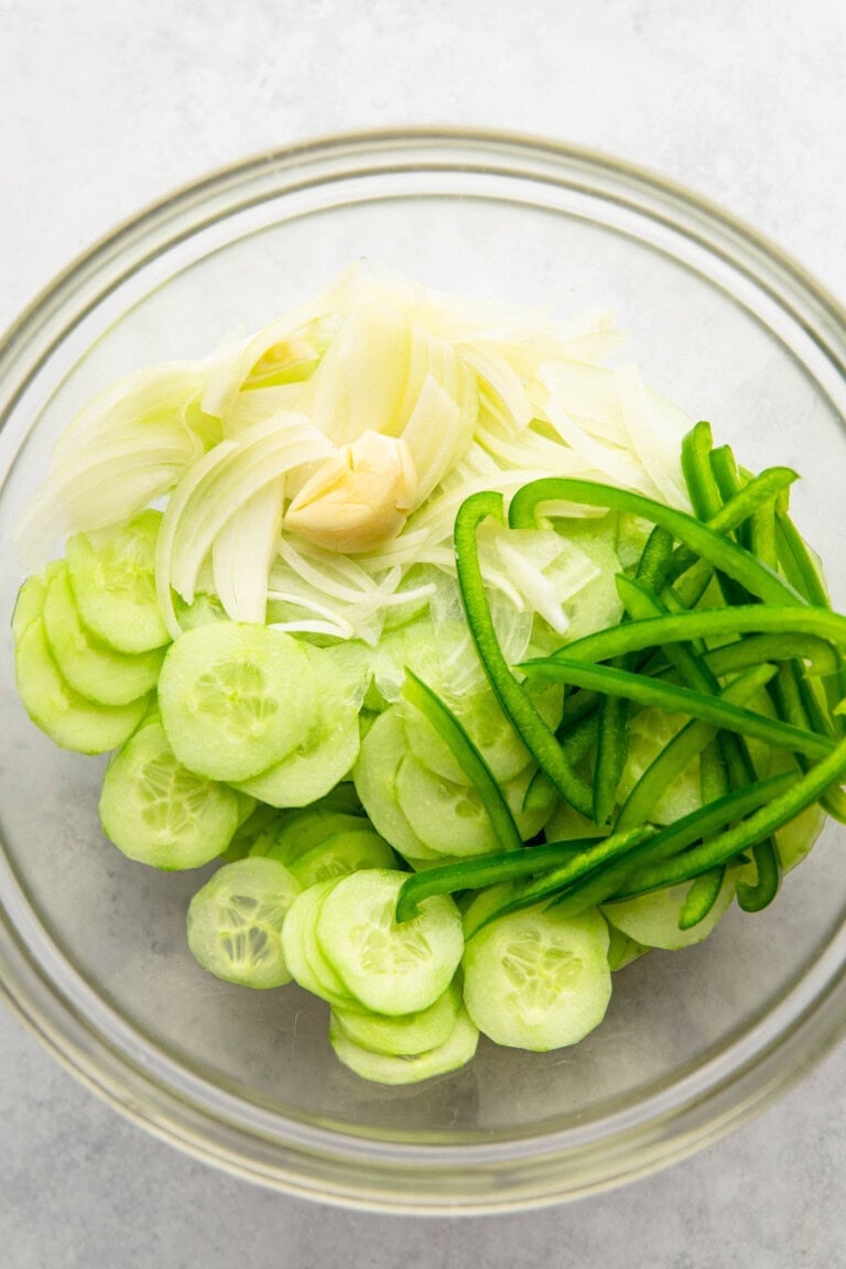 A glass bowl containing sliced cucumbers, thinly sliced onions, a clove of garlic, and thinly sliced green bell peppers, placed on a light-colored surface.