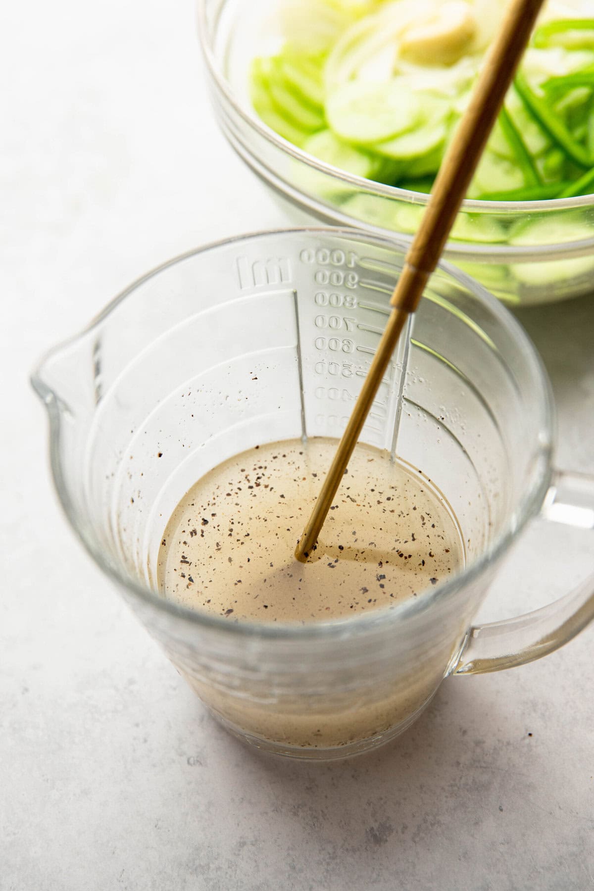 A measuring cup filled with a seasoned liquid, stirred with chopsticks. A bowl with sliced green vegetables is visible in the background.