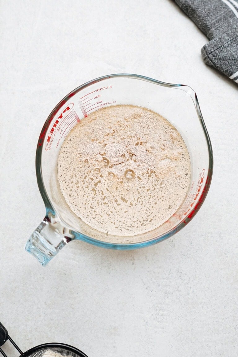 A glass measuring cup filled with a bubbly liquid mixture sits on a light-colored surface, with a dark gray cloth partially visible in the top right corner.