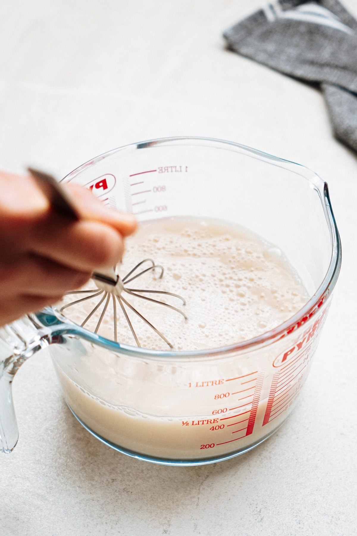 A hand whisks a frothy liquid in a clear measuring cup on a kitchen counter.