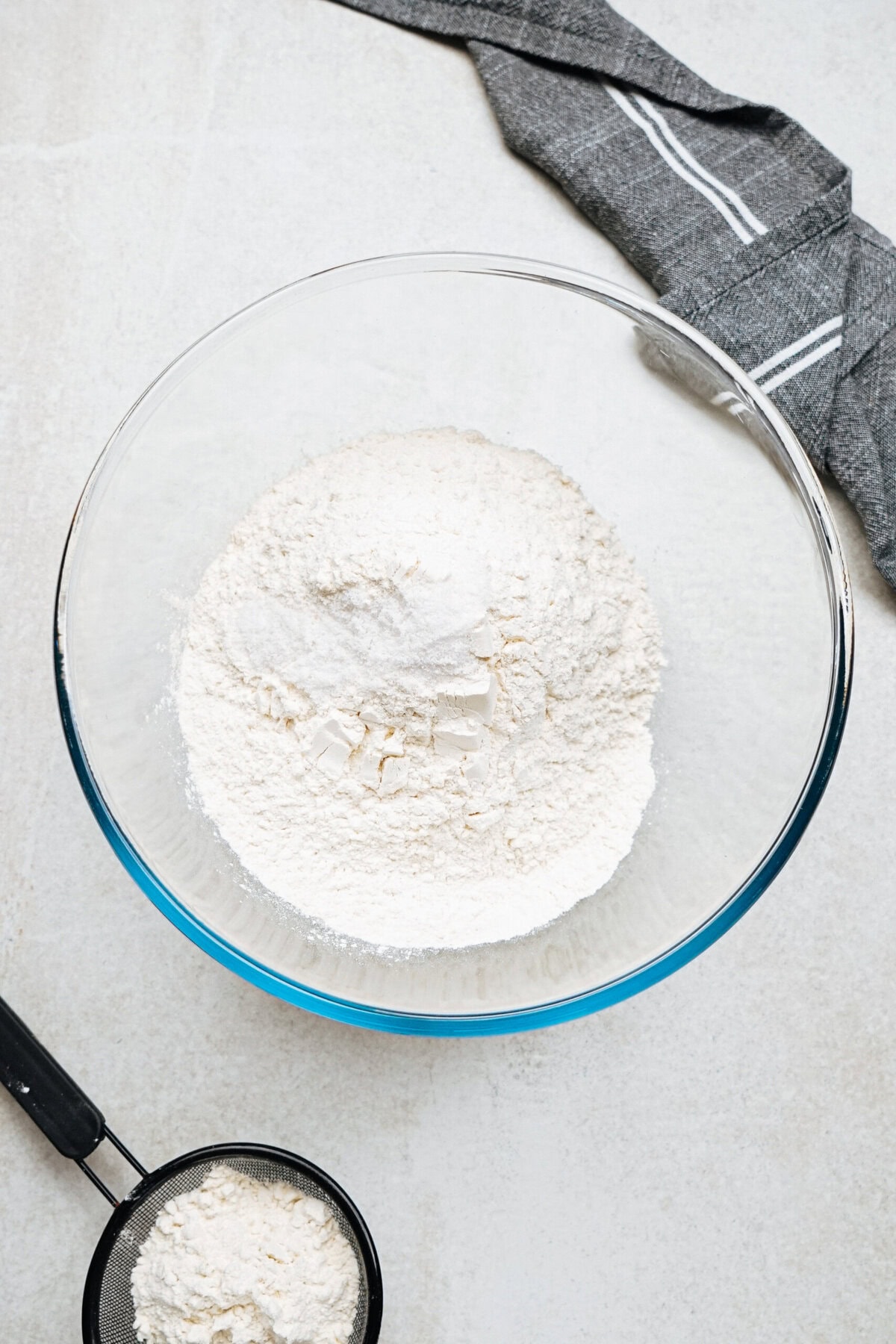 A glass bowl containing flour is placed on a light surface with a small sifter and a gray cloth nearby.