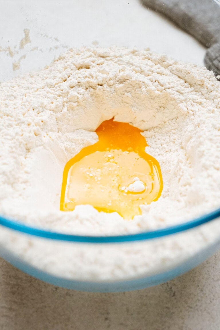 A glass bowl filled with flour has a well in the center containing a cracked egg yolk.