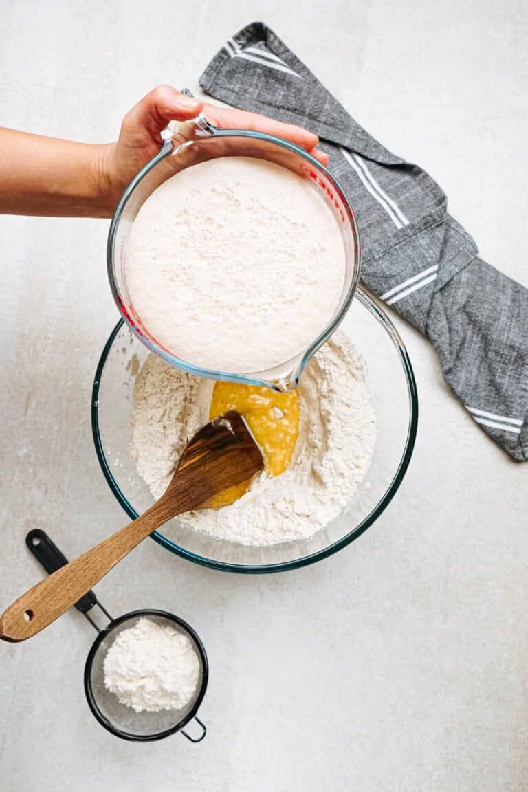 A hand pours a milky substance from a measuring cup into a bowl of flour and melted butter with a wooden spoon, next to a small sifter and a gray cloth on a light countertop.