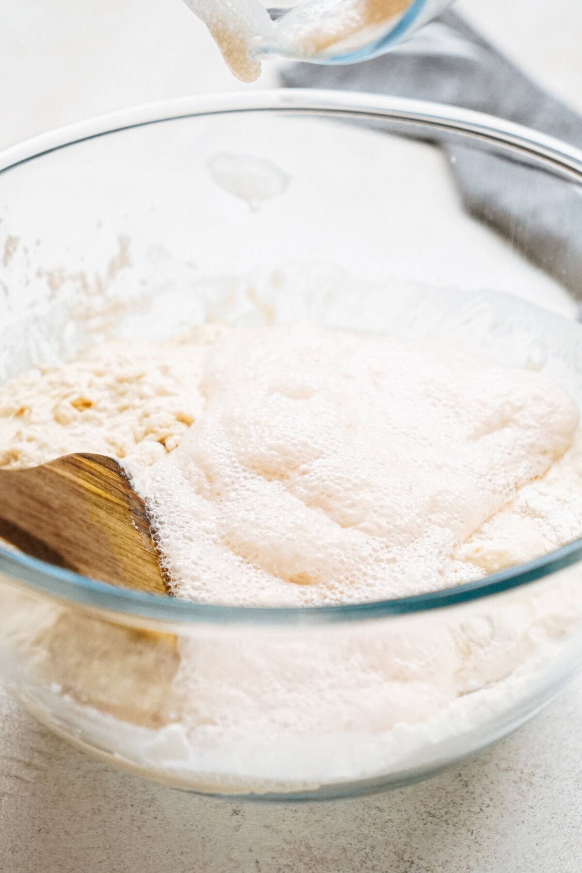 A wooden spoon mixing frothy yeast mixture and flour in a glass bowl.