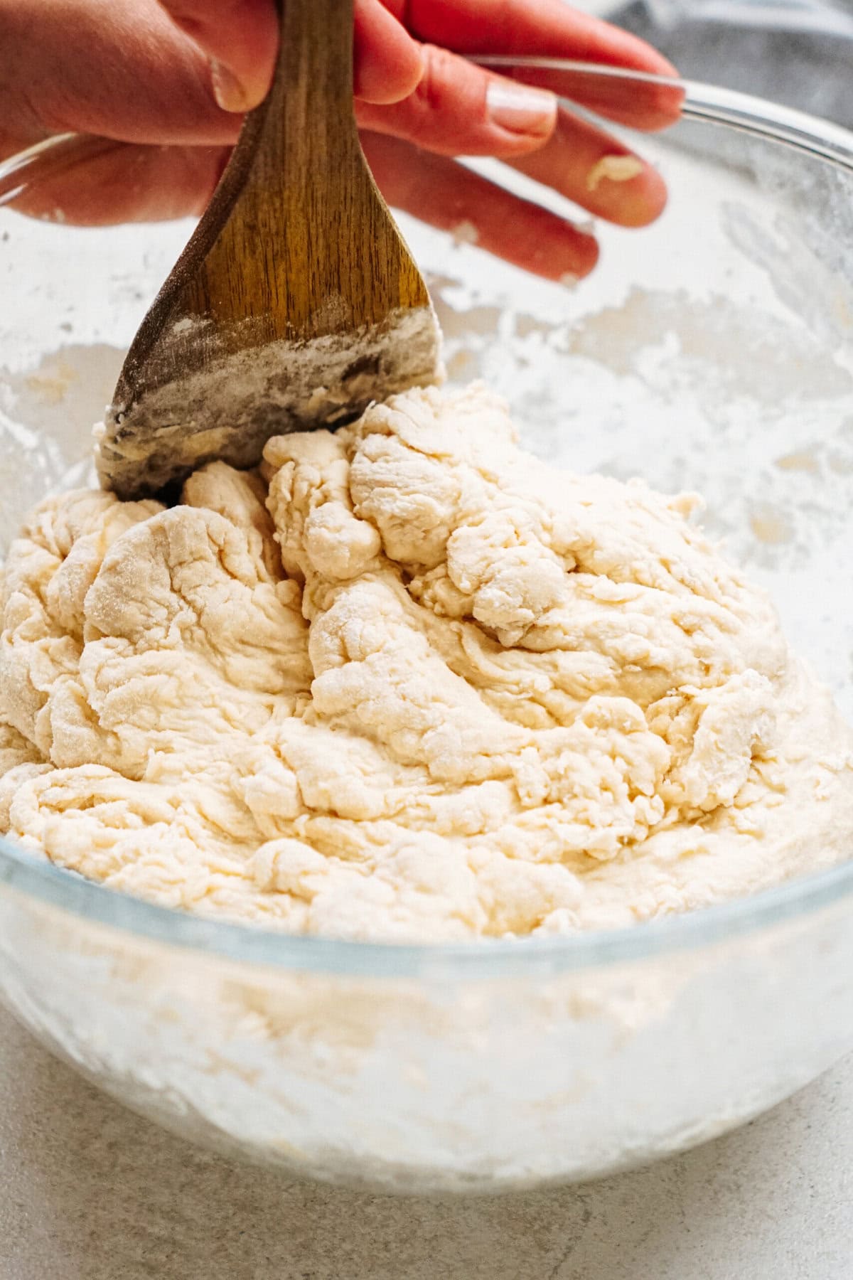 A wooden spoon mixing dough in a glass bowl. Hands are visible holding the spoon.