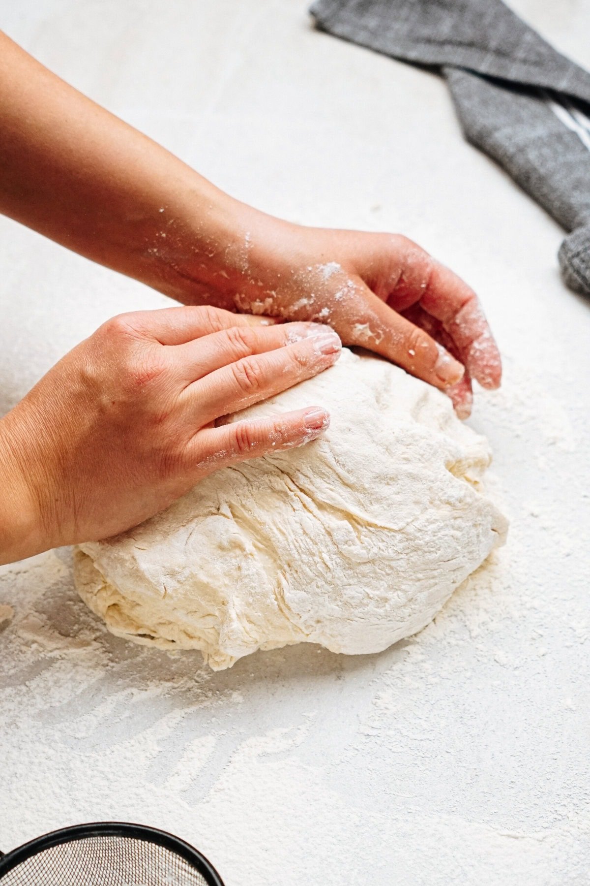 Hands kneading a ball of dough on a floured surface with a gray cloth nearby.